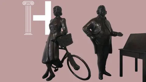 Harris Museum The dark bronze sculptures of suffragette Edith Rigby and industrialist Sir Richard Arkwright against a dull pink background. Rigby is leaning with her back against a bicycle and Arkwright is standing by a desk pointing with his left hand as his right rests on his hip next to a round tummy