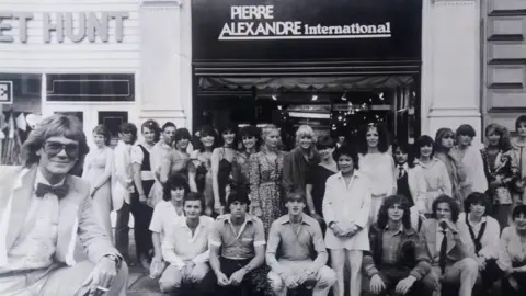 Family photograph A black and white image shows Pierre Alexandre, leaning on his knee with a cigarette in his hands, is wearing black glasses and a suit with dicky bow. He is standing in front of a crowd of people outside of a shop bearing the words 'Pierre Alexandre International'