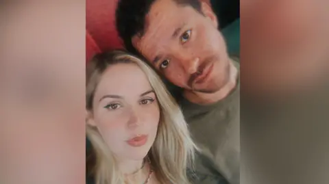 A woman with long blonde hair is looking at the camera. She is next to a man with brown hair and a moustache.