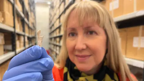 Leeds City Council A blonde woman wearing an orange jumper, black and yellow scarf and blue rubber gloves holds a small piece of animal bone carved into a hook shape. There are shelves of archive boxes behind her.