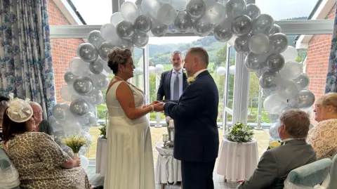 BBC A photo of Rachel Berry, and her new husband, Mike Berry, at the top of the alter surrounded by guests during the wedding ceremony. Rachel wears a white long wedding gown, has a short haired, curly updo, and holds hands with Mike, who wears a navy suit. The celebrant stands inside two patio doors, wearing a suit. A silver balloon arch is behind the couple and over the head of the registrar. 