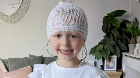 Noa-Rose smiling looking at the camera. She wears a white t-shirt and has a bandage around her head with colourful wires coming out at the back. 