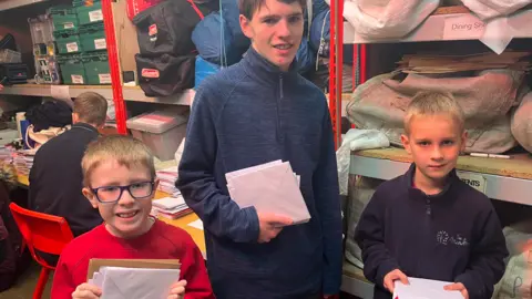 Wayne Bavin/BBC Three young boys standing together in a sorting office holding piles of Christmas cards in envelopes. On the left, a boy with fair hair and black-rimmed glasses wearing a red jumper. In the middle, an older boy who is much taller in a blue fleece. On the right, a shorter and young boy, wearing a navy-blue or purple fleece.