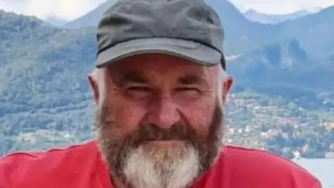 An Garda Síochána A man, wearing a red top, pictured from the shoulders up. He is wearing a hat. He has a grey beard.