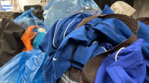 Close-up of a pile of blue clothing or uniforms draped over a container, surrounded by large blue plastic bags filled with items. The scene appears indoors with chairs and shelves in the background.