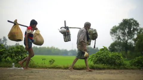 BBC Two generations of a Rohingya refugee family walk with their belongings attached to long sticks across their shoulders