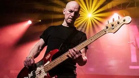 A bald man wearing a black shirt plays a red and white bass guitar on stage. Yellow and red lights are visible behind him.