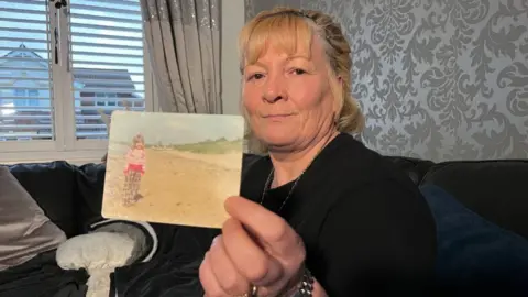 Michelle Kilpatrick, who has blonde hair tied back, holds a photo of herself as a child up to the camera while sitting on a sofa. In the photo she is standing on a beach with a blue sky in the background. 