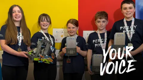 Five primary pupils, three girls and two boys, smiling at the camera, with a robot and trophies.