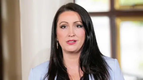 Deputy First Minister Emma Little-Pengelly has long black hair and brown eyes. She is wearing a purple suit and a black top.