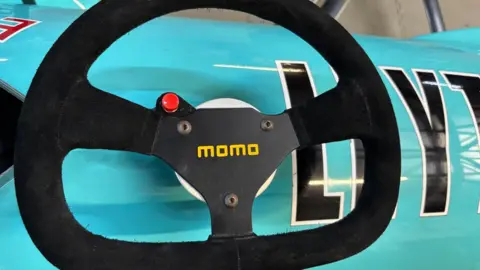 A black steering wheel with a red button and yellow font saying "momo" on it. The wheel is placed on a turquoise car.