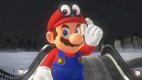 Tangkapan Layar Nintendo menunjukkan Mario di dunia dari Super Mario Odyssey, menatap penonton dengan penuh teka-teki saat ia menyesuaikan pinggiran topi merahnya yang terkenal dengan tangan bersarung tangan putih.