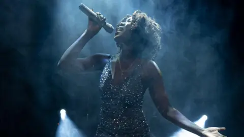 Queen of the Night A woman playing Whitney Houston in a show called Queen of the Night at the Bristol Hippodrome. She is wearing a sparkling dress and is holding a microphone above her head as she sings