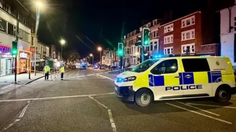 Leicester Media Online A police van in front of police tape along a road with no traffic on.