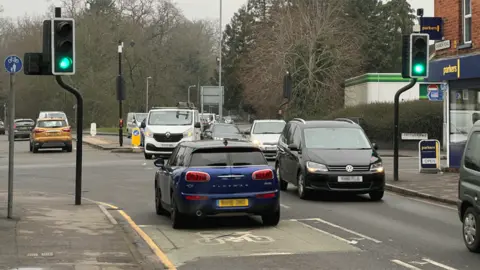 A crossroads full of traffic coming to and fro. The traffic lights are green. A BP can be glimpsed on the far right corner.