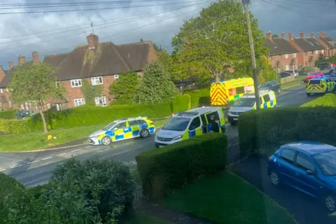 Other Four West Mercia Police vehicles are parked in a road. Green hedges and trees can be seen lining the road. One ambulance is also parked. 