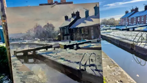 Rob Pointon A painting of a canal lock, in place in front of the actual scene depicted. In both the painting and photo, a row of terraced houses are visible on the other side of the canal. 