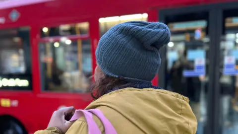 A woman in a coat stands facing away from the camera next to a London bus