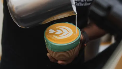 BBC A barista crafting a milk foam design on top of a freshly brewed coffee in a mug