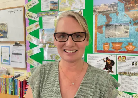 Gemma Dawson/BBC Teacher Esther Skelland. She has tied back blonde hair and dark-rimmed glasses and is standing in a classroom. There are books on a shelf behind her.