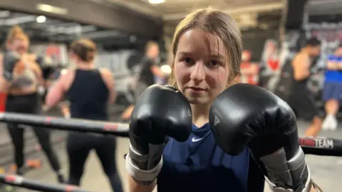 A 19 year-old woman is standing next to a boxing ring. She is wearing black boxing gloves which she is holding up close to her face. She has light brown hair tied back and is wearing a navy T-shirt. Other poeple are training in the ring behind her.