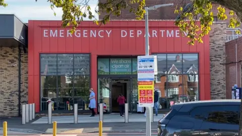 Anne-Marie Tasker/BBC An exterior view of the entrance to the emergency department at Scunthorpe General Hospital. The glass-fronted entrance is red with the words "Emergency department" above the door. There is a staff member in uniform exiting the building.