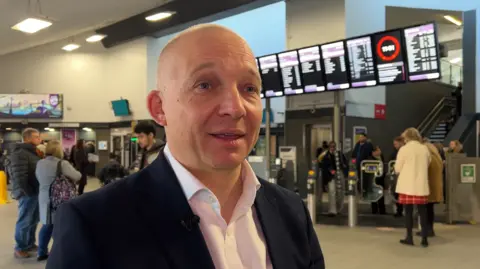 Will Rogers, un hombre blanco y calvo, aparece en la foto con una chaqueta oscura en el vestíbulo de una estación de tren.