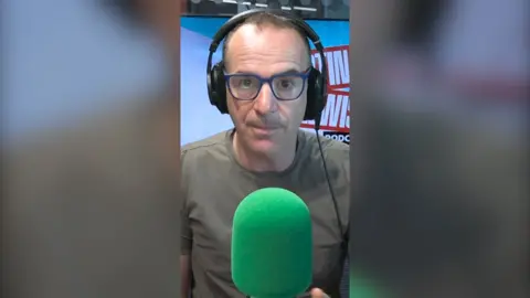 Martin Lewis is wearing dark green T-shirt and black-framed glasses. He is wearing headphones and sitting behind a green mic.
