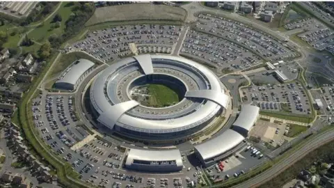 An aerial view of GCHQ headquarters, a circular, silver metal coloured building with several small units nearby, surrounded by car parks, a public road and a housing estate.