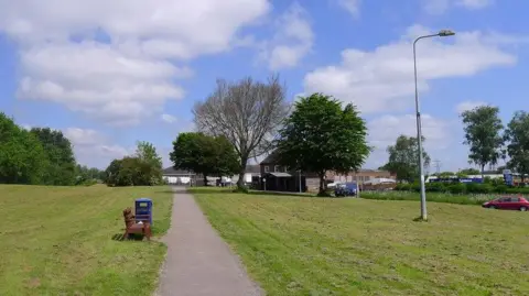 Tim Heaton/Geograph A park with lots of greenery, a lamppost, a wooden bench and a bin. A concrete pathway runs through the park.