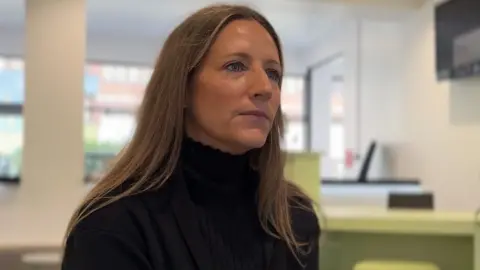 A woman with long blonde hair looks at someone off camera. She is wearing a black polo neck and black blazer and in a white-walled room with a green desk and windows behind her.