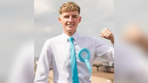 A young man with curly blond hair which is short at the sides is wearing a long-sleeved white shirt and a turquoise blue tie with a turquoise Reform UK badge. He is smiling and holding one fist up in the air.