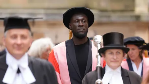 Stormzy is in the middle of a crowd, which is blurred, walking towards the camera. He is wearing a black T-shirt and suit jacket, and has a flat doctorate hat on and a pink and red gown. 