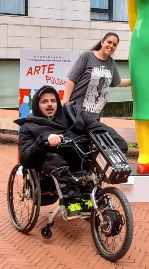 Israel Vidal A man in a black hoodie is sitting in a wheelchair. The wheelchair has an electric wheel attachment.