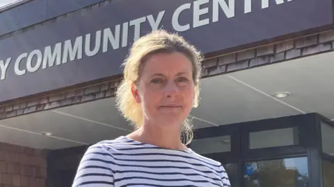 Caroline Speca stands in front of a community centre entrance and smiles at camera, she has blonde hair and is wearing a striped shirt