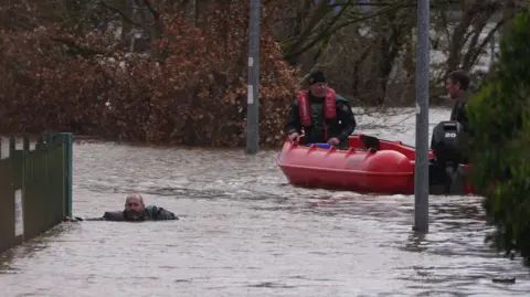 Niall Carson/PA Wire Members of Slaney Search and Rescue working in floodwater in Enniscorthy. Two meb are sitting in a red dinghy while another man is swimming in the flood up to his chest. He is holding on to a green fence. 