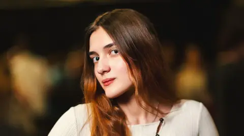 BBC/Julia Moore Young woman with long brown hair and beige top with turned head looking into the camera. Blurred background.