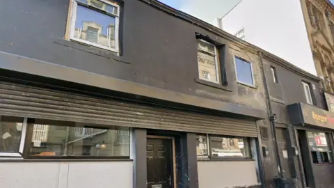 A dark-painted two-storey building on a narrow street, with a takeaway entrance at ground level and windows on the upper floor.