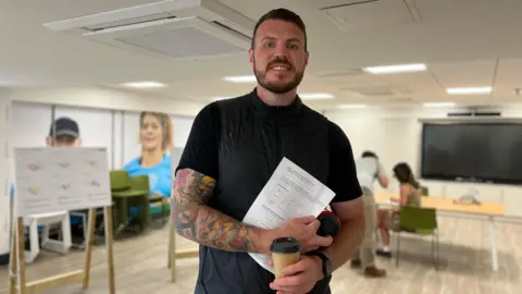 Nick has tattooed arm stands in the foreground holding a coffee cup and papers. In the background, several people are seated at tables or viewing posters on easels in a modern, well-lit room with a large screen on the wall. All faces are blurred for privacy