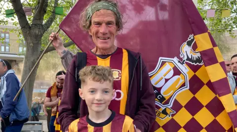 A man in his 60s with hands on boys shoulders wearing Bradford City tops smiling with flag behind them