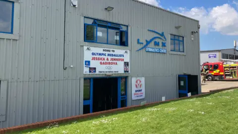Alex Pope/BBC The exterior of a grey metal building with blue-framed windows and doors. A banner on the wall says “Home to Olympic medallists Jessica & Jennifer Gadirova – Tokyo Olympics 2020,” with photos printed on it and Olympic rings. 