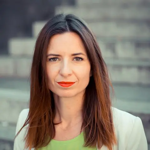 Franziska Weir A woman with long dark hair and bright red lipstick in a green t-shirt and a lighter green jacket