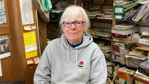 A phot showing a woman sat down in front of a bookcase. She has grey hair and is wearing glasses and a a grey hooded jumper