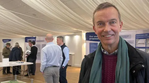 Connor Bennett/BBC A man smiles at the camera. He has short brown hair and blue eyes. He is wearing a green scarf, burgundy jumper and a black jacket. Behind him are people standing and talking to each other in two groups, with information boards around the perimeter of the room. 