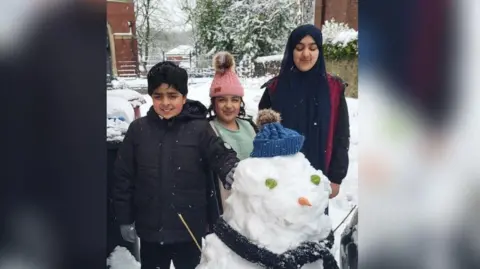 IqbalKhadijah A boy and two girls stand next to their snowman, who has a carrot nose and sprouts for eyes, while wearing a blue bobble hat.