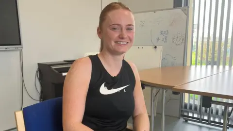 Nicola Fagan sits in a blue chair beside a wooden-topped table. She is wearing a black sleeveless sports top with a white Nike swoosh logo on the front. She has auburn hair pulled back in a ponytail.