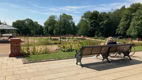 A general view of a park which has a bandstand in the middle surrounded by pathways and flowers, with trees in the distance. A woman is sitting on a bench on the right, next to a bicycle. 
