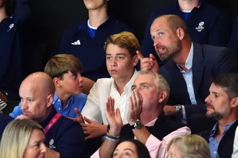 PA Media Prince William leans in to speak to his sons George and Louis while the family watch a netball match. William points to something above his eyeline while Louis' attention is on his father. George stares straight ahead.
