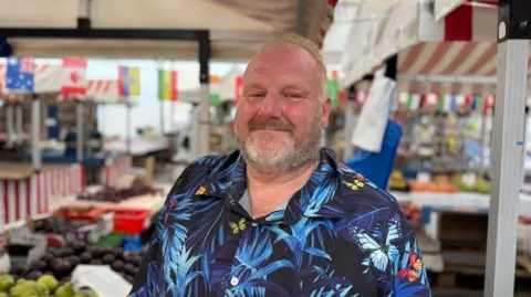 A bearded man in a flowery shirt standing in front of a market stall