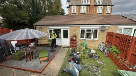 Steve Hubbard/BBC Back garden of a bungalow with green grass and garden furniture, with a police man dressed in his uniform, standing by the back door.